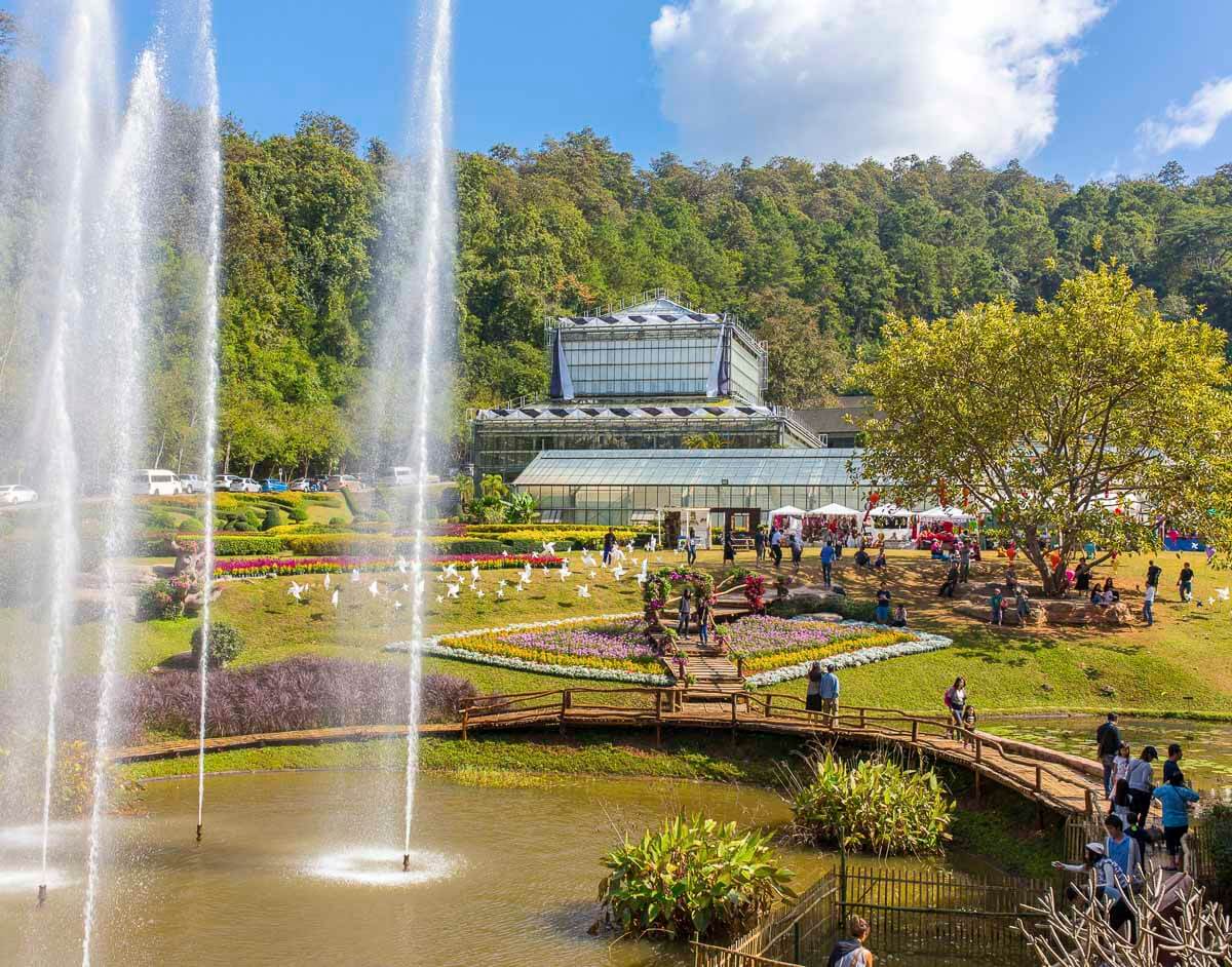 O Jardim Botânico Rainha Sirikit, com fontes de água e estufa de vidro, rodeado por vegetação e visitantes explorando a área.