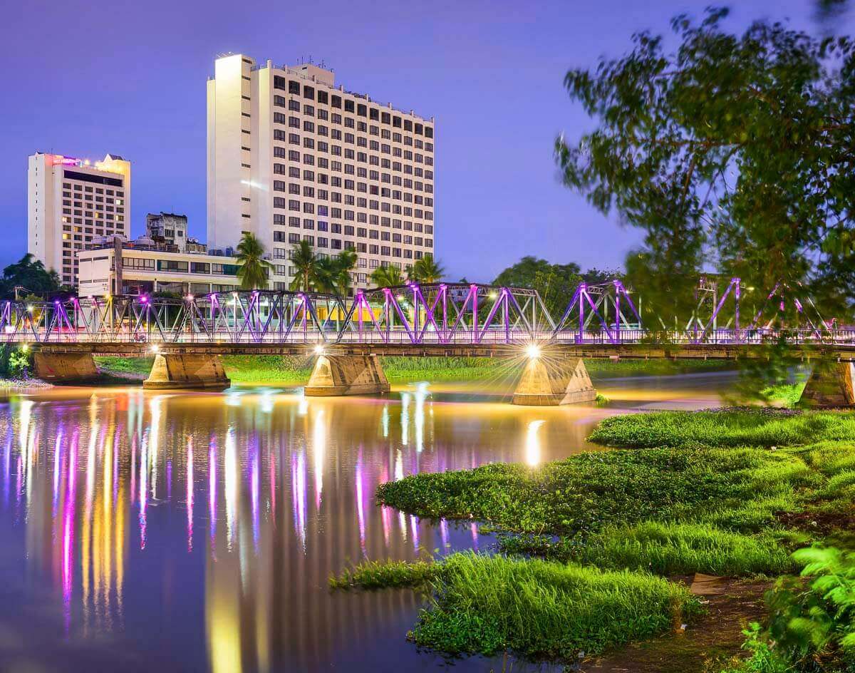Ponte iluminada sobre o rio Ping em Chiang Mai, refletindo luzes coloridas na água durante a noite.