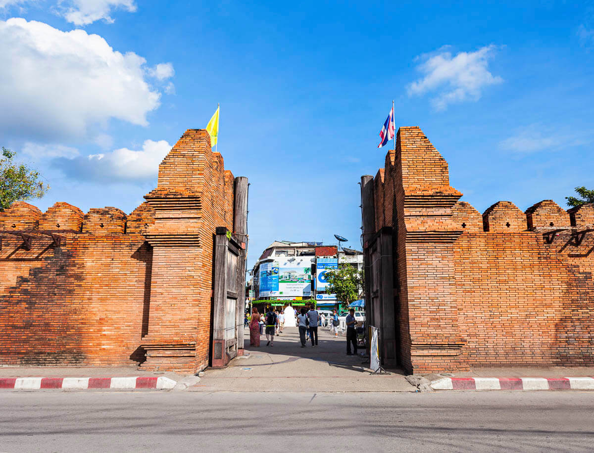 Portão de tijolos vermelhos em Chiang Mai, com bandeiras tailandesas e pessoas passando ao fundo.