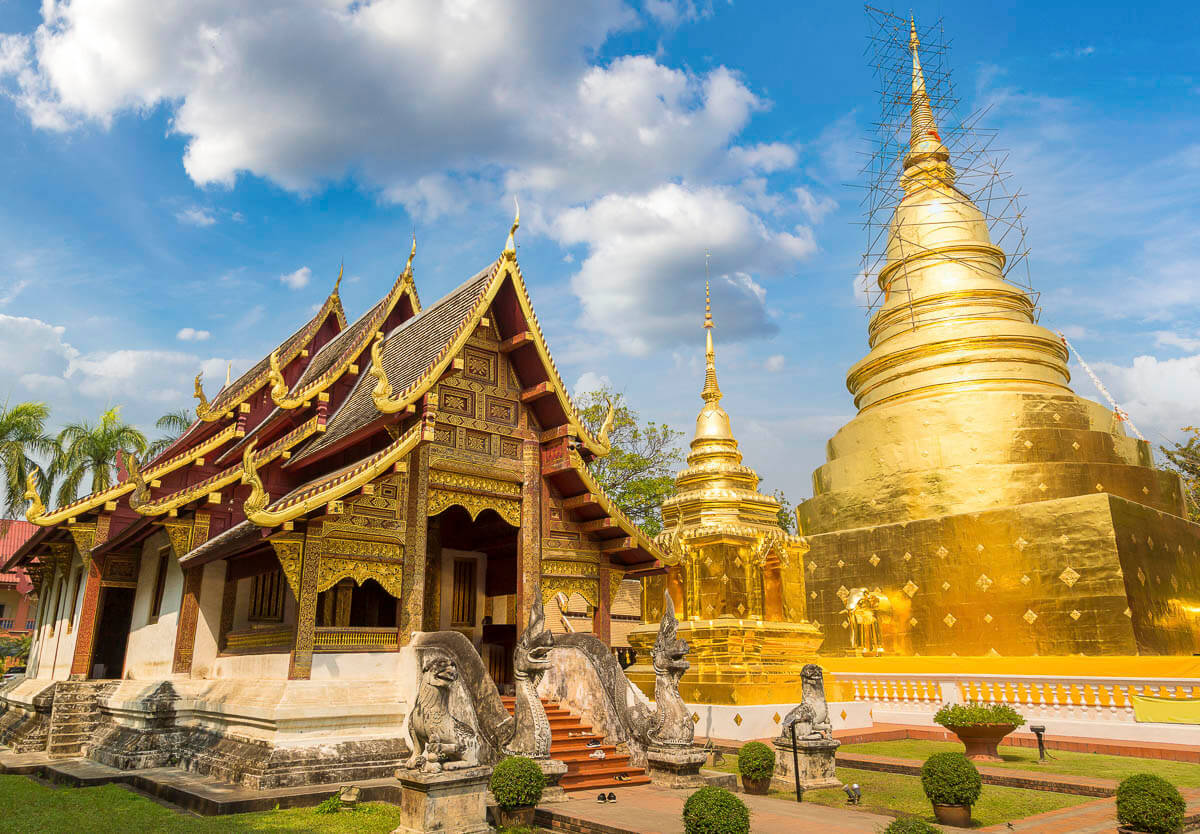 Templo Wat Phra Singh em Chiang Mai, com uma pagoda dourada brilhante ao lado de uma estrutura de madeira com ornamentos detalhados, cercado por esculturas e jardins.