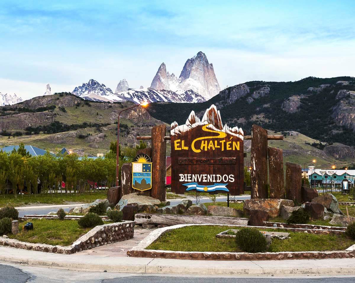 Placa de boas-vindas na entrada de El Chaltén com vista para o Fitz Roy ao fundo, montanhas nevadas e vegetação baixa.