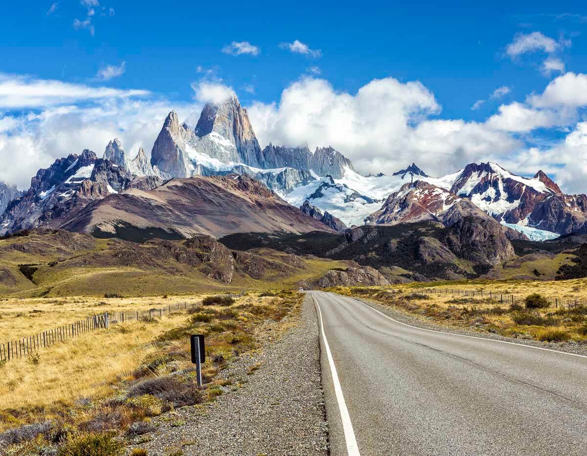 Estrada reta cercada por campos abertos que leva ao Fitz Roy, com montanhas nevadas e céu azul ao fundo.