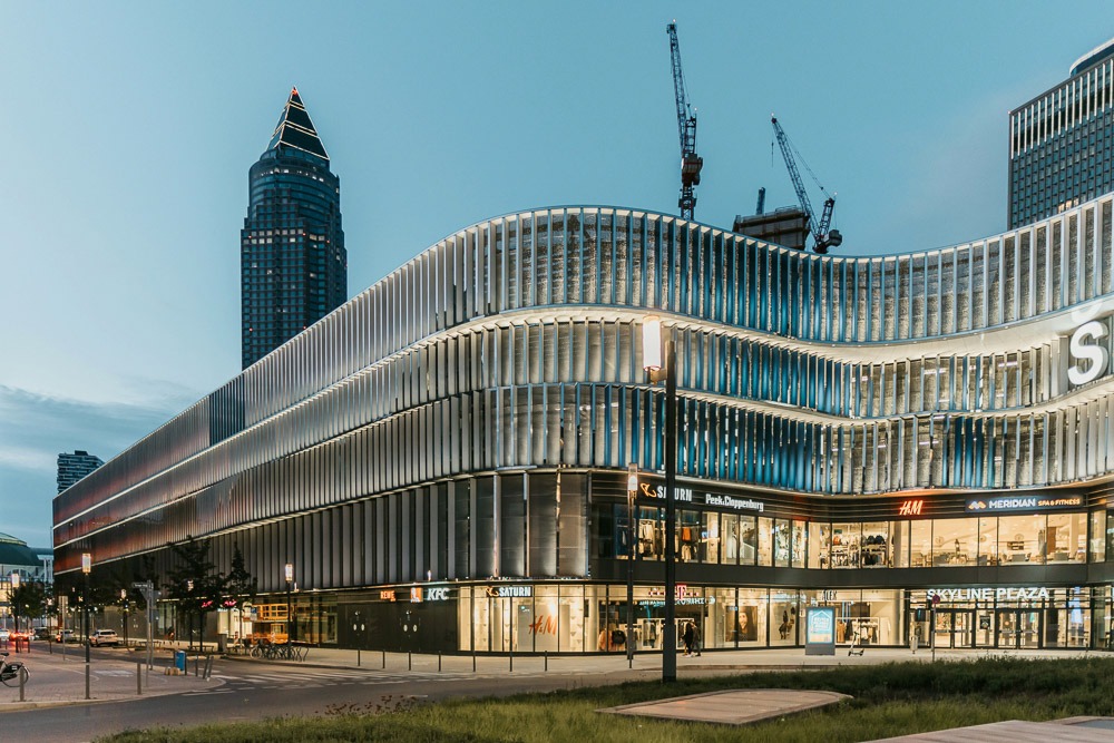 Centro comercial Skyline Plaza em Frankfurt, com arquitetura moderna iluminada, ao lado do distrito Gallusviertel, visto ao entardecer. No fundo, o arranha-céu Messeturm e guindastes de construção.