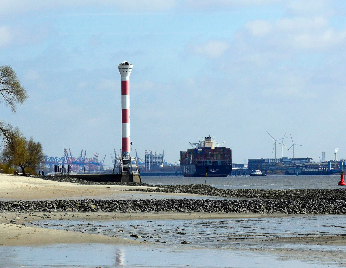 Farol vermelho e branco na Elbstrand Altona, Hamburgo, com o porto ao fundo e praia na frente.