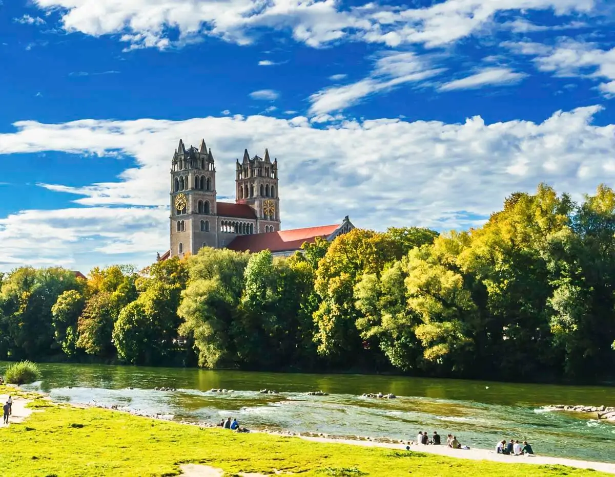 Igreja de São Lucas em Munique, cercada por árvores e localizada às margens do Rio Isar no parque Flaucher, sob um céu azul brilhante.