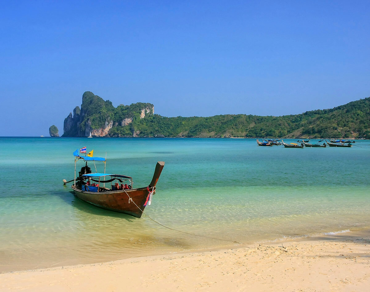 Praia de Loh Dalum com um barco tradicional tailandês ancorado na água clara e colinas verdes ao fundo.
