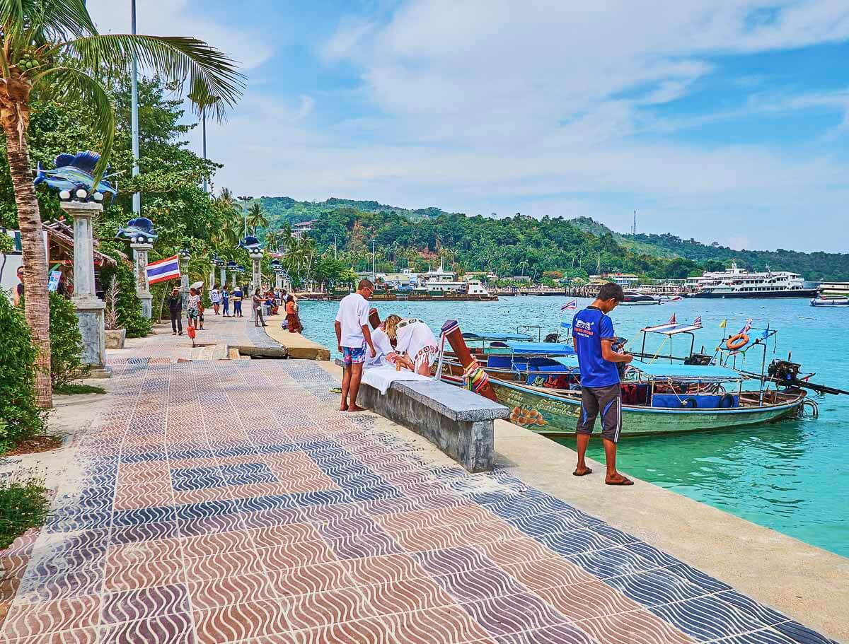 Calçadão à beira-mar no oeste de Tonsai, com palmeiras, barcos e pessoas passeando.