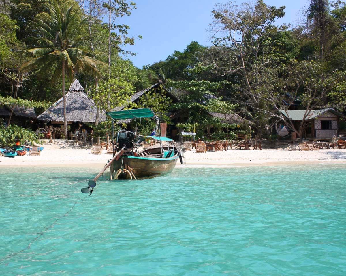 Praia isolada em Viking Beach com um barco tailandês na água e cabanas cercadas por vegetação.