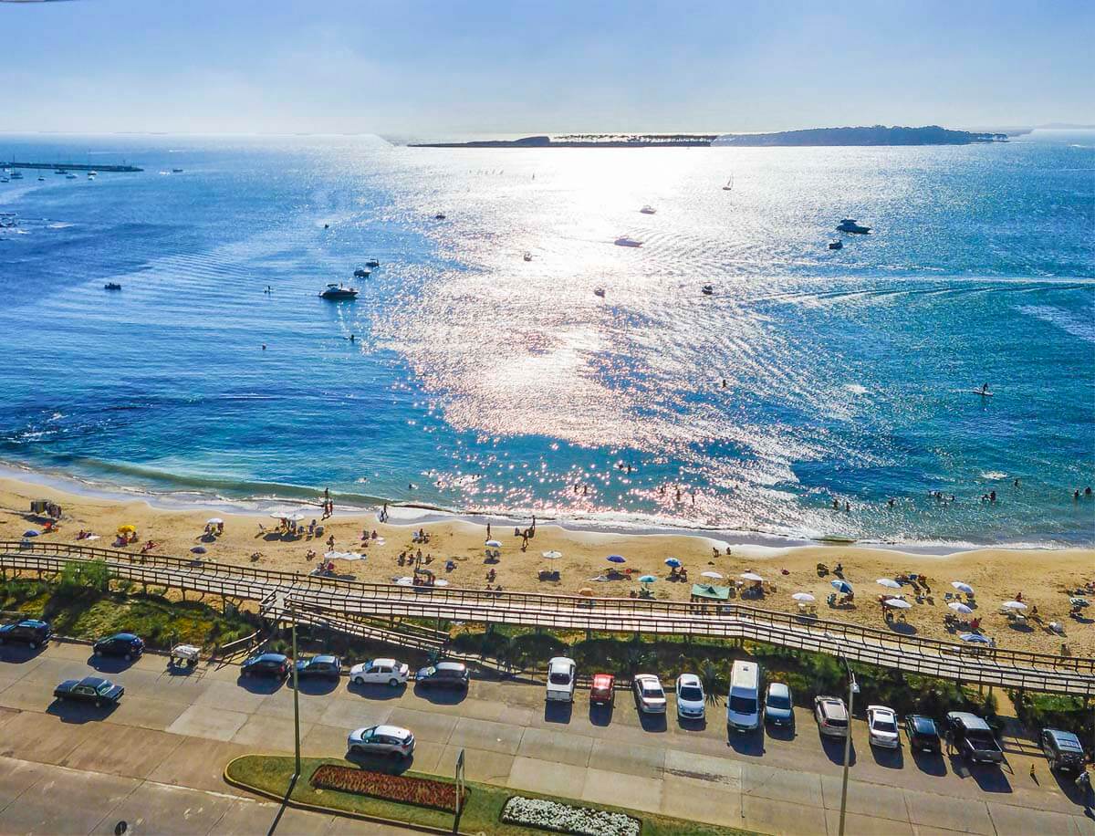 Vista da Praia Mansa com areia dourada, guarda-sóis e pessoas aproveitando as águas calmas sob o céu ensolarado.