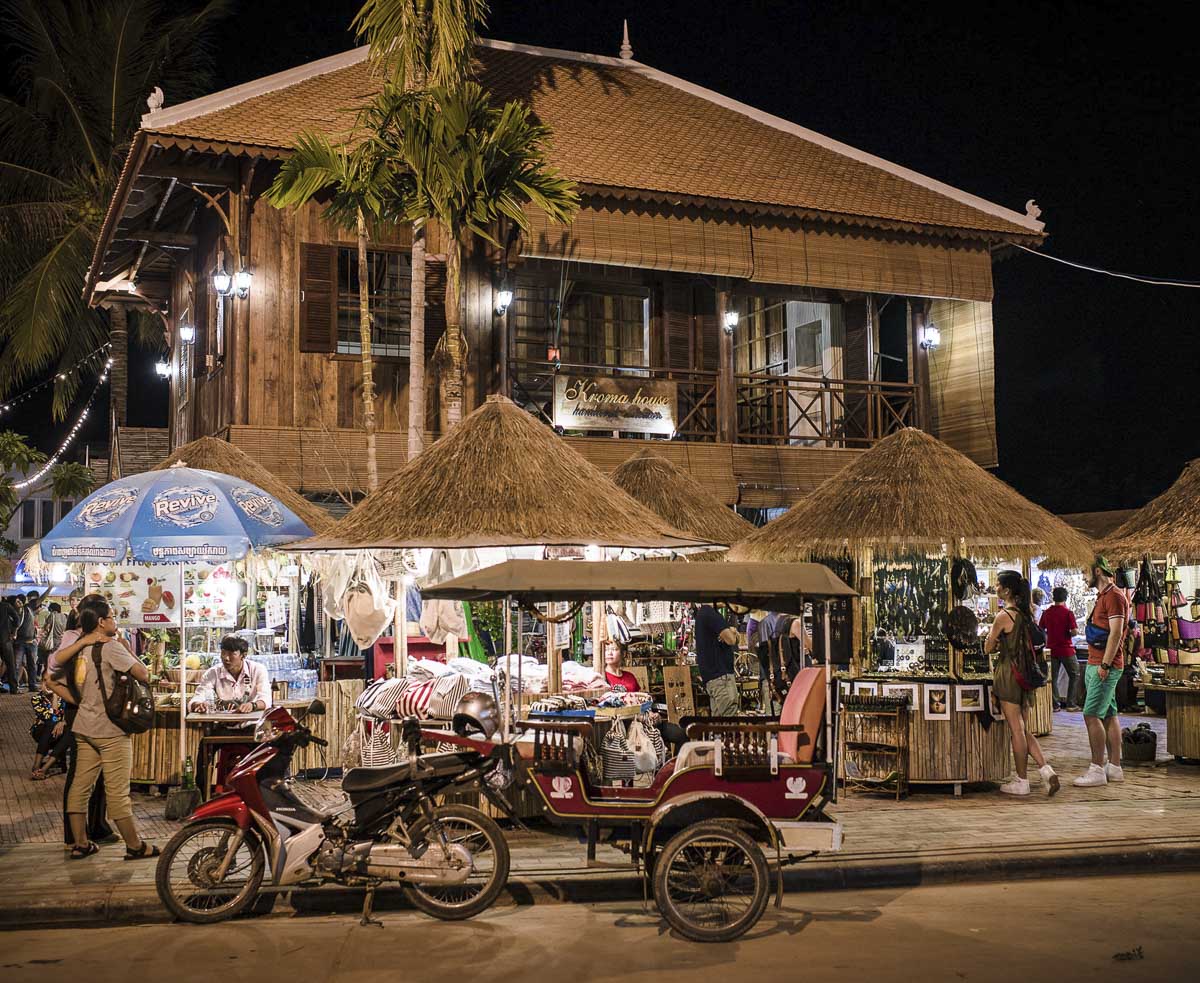 Fachada do mercado noturno de Angkor em Siem Reap, com barracas de palha iluminadas, tuk-tuk estacionado e pessoas explorando os produtos à venda, em frente a um prédio de madeira com varanda.