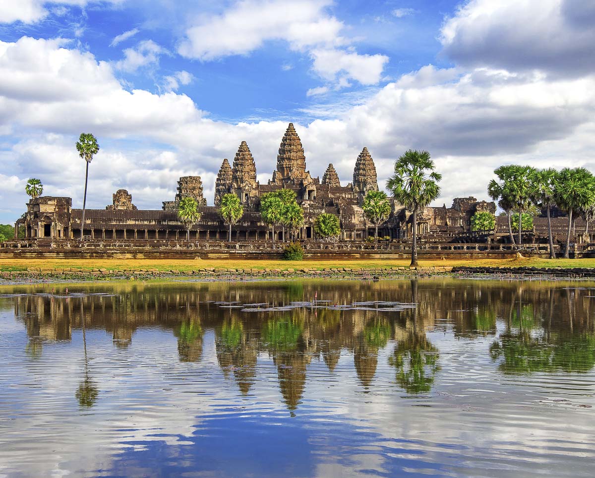 Templo Angkor Wat refletido em um lago, cercado por palmeiras e vegetação sob um céu parcialmente nublado.