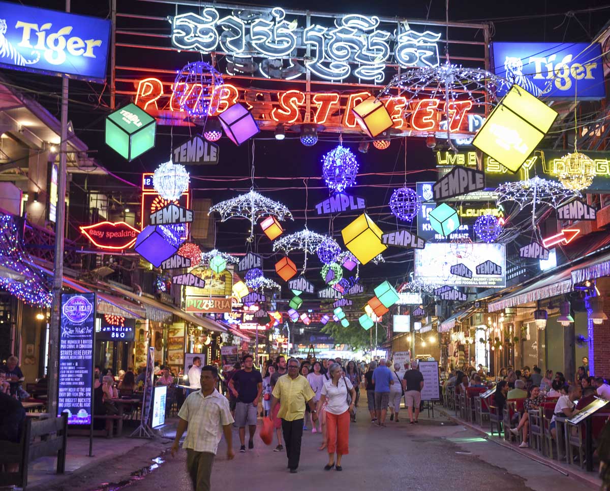 Rua Pub Street em Siem Reap iluminada por letreiros neon coloridos e decoração vibrante, com pessoas caminhando entre restaurantes e bares.