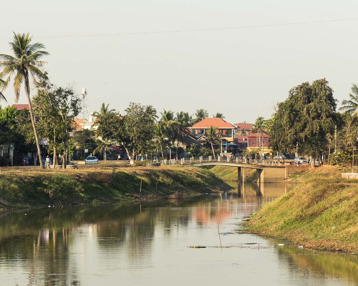 Ponte atravessando o rio em Siem Reap, com vegetação nas margens e casas ao fundo sob um céu claro.