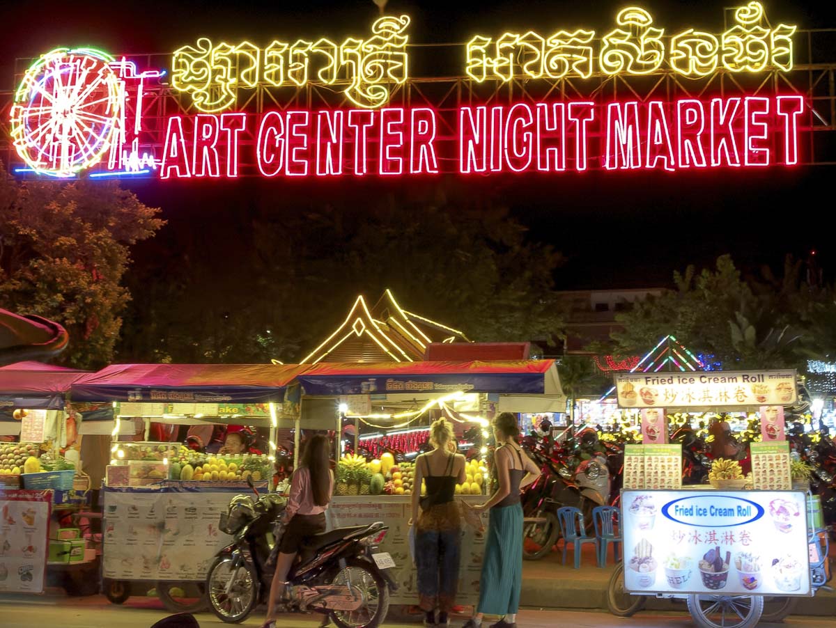 Entrada iluminada do Art Center Night Market em Siem Reap, com letreiro neon vermelho e barracas coloridas de alimentos e mercadorias.