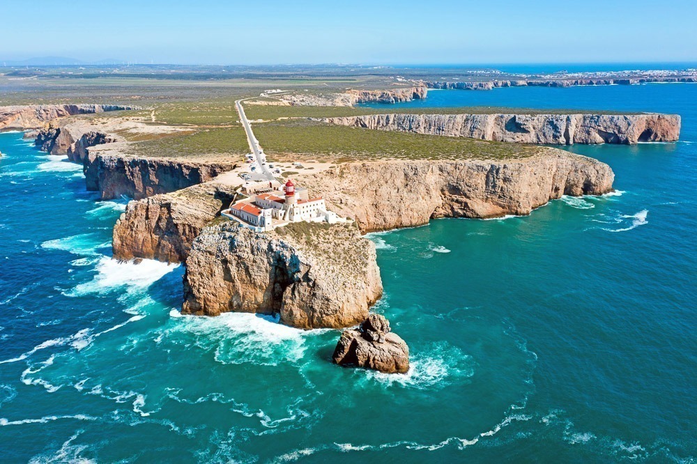 Vista aérea do Cabo de São Vicente, em Sagres, Algarve, com falésias imponentes e um farol no topo de uma formação rochosa cercada pelo oceano azul profundo.