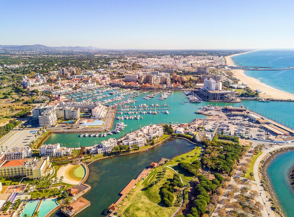 Vista aérea de Vilamoura, no Algarve, destacando a marina repleta de barcos e iates, hotéis luxuosos e praias douradas à beira do oceano Atlântico.