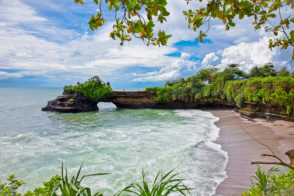Vista da praia Batu Bolong em Canggu, Bali, com ondas quebrando suavemente na areia, cercada por vegetação e céu parcialmente nublado.