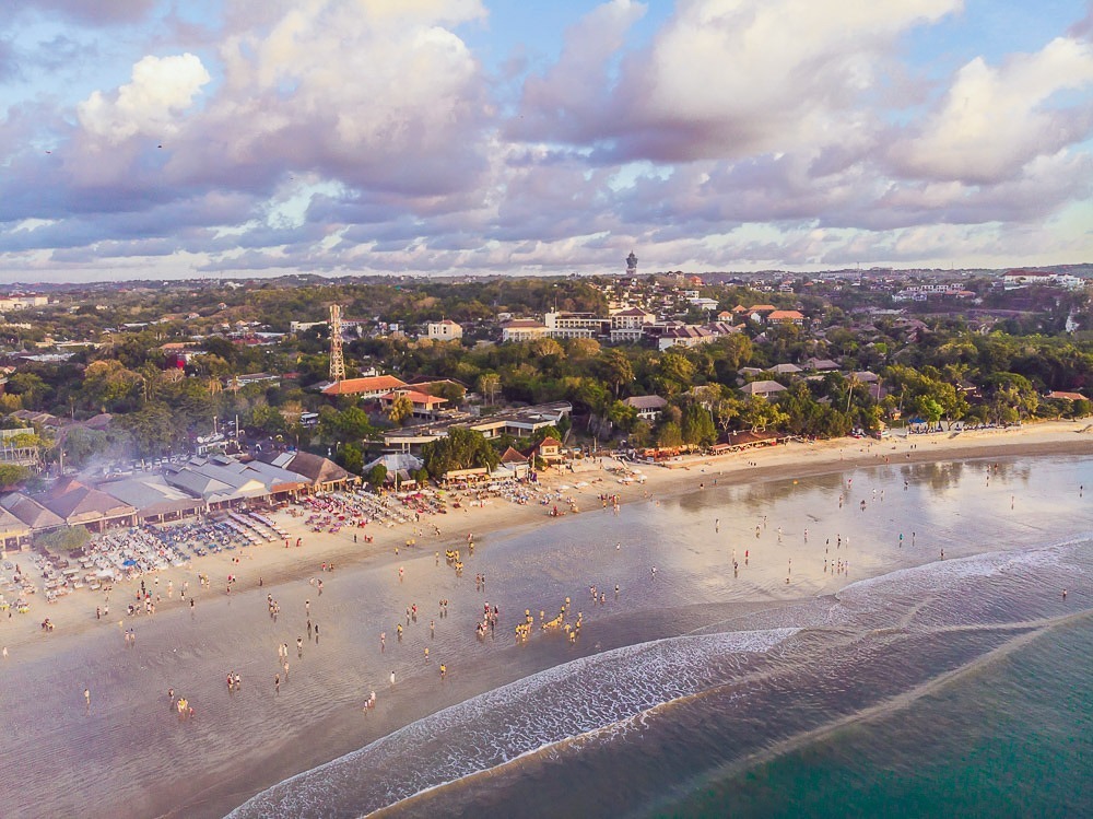 Vista aérea da praia de Jimbaran em Bali, com uma linha de restaurantes e pessoas aproveitando o pôr do sol na areia.