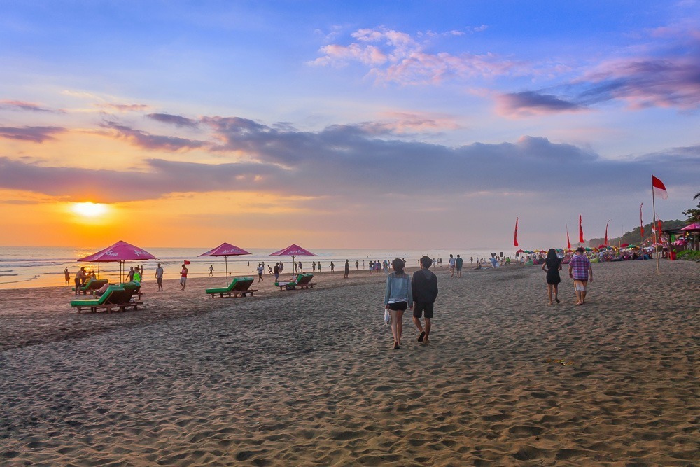 Praia de Legian ao entardecer, com guarda-sóis rosa e pessoas passeando na areia.