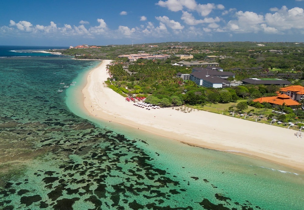 Vista aérea da praia de Nusa Dua, com areia branca, águas cristalinas e resorts ao fundo.