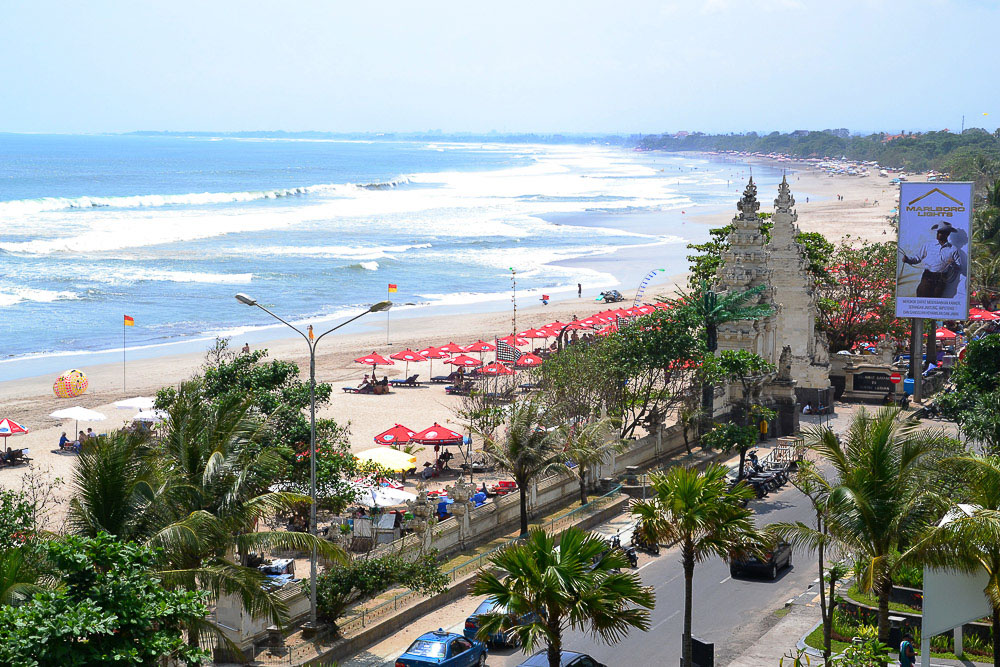 Vista de Kuta Beach em Bali, com ondas quebrando na areia, guarda-sóis vermelhos e uma estrada ladeada por palmeiras ao fundo.