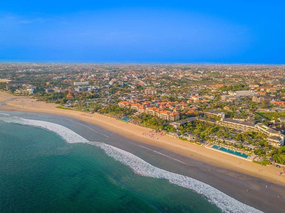 Vista aérea de Seminyak, Bali, com uma longa praia de areia dourada, resorts luxuosos e o mar azul profundo.