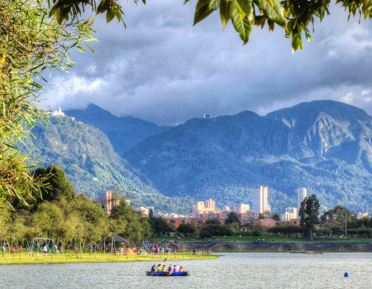 Parque Simón Bolívar em Bogotá, com lago, árvores e montanhas ao fundo. Pessoas aproveitam o espaço em barcos e áreas gramadas.