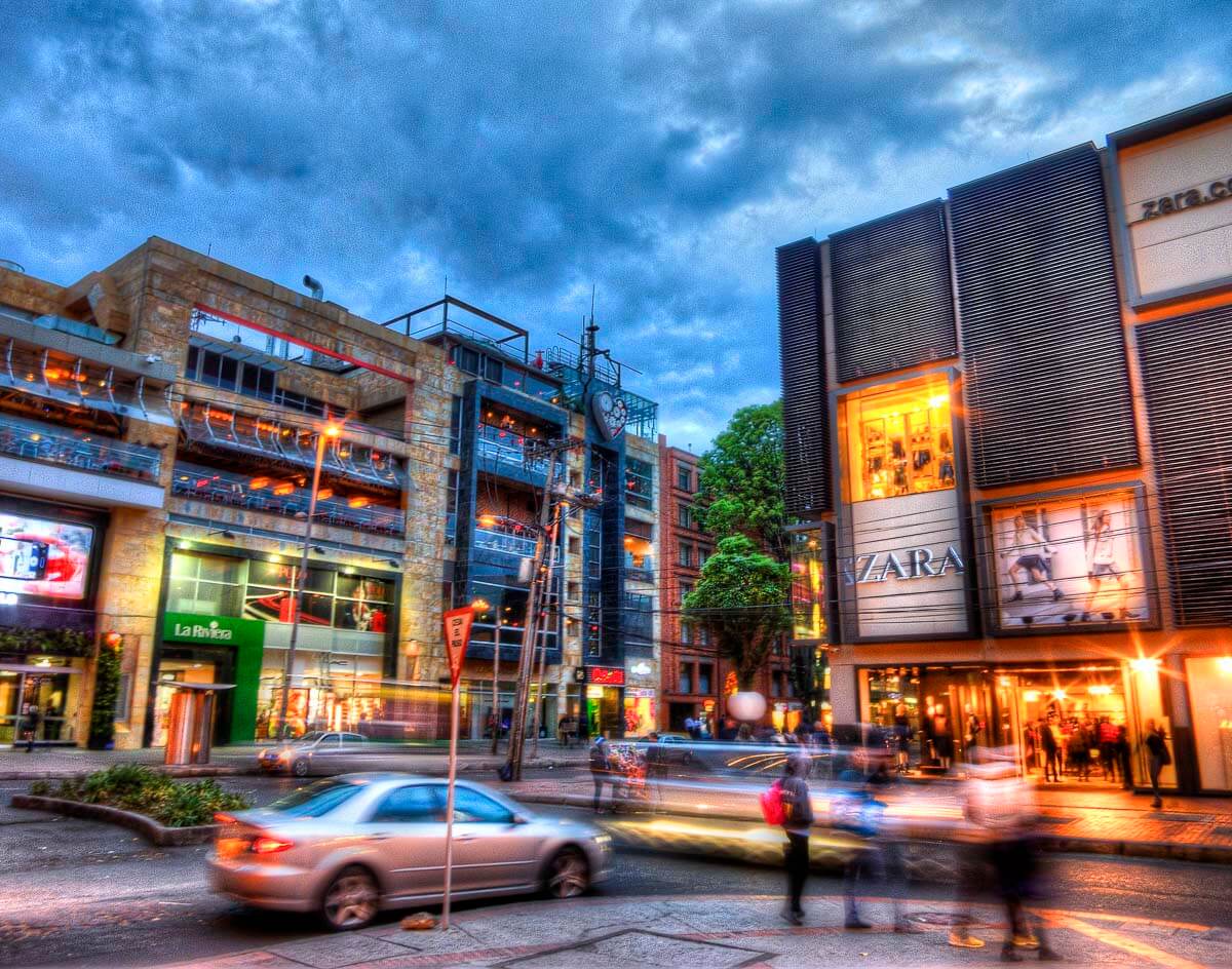 Rua movimentada na Zona Rosa de Bogotá, com lojas de luxo e prédios modernos iluminados ao entardecer.