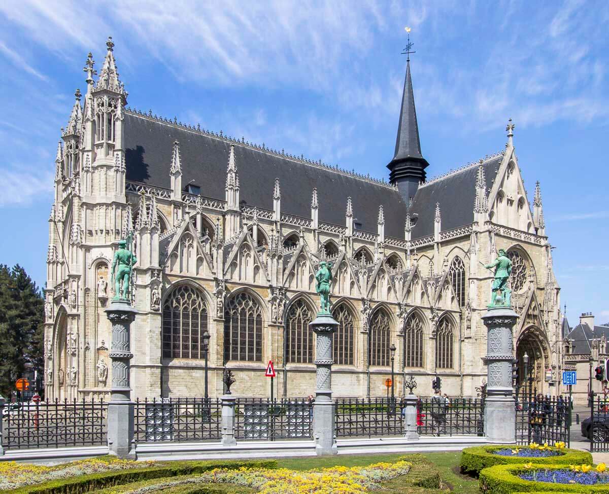 A Igreja de Notre-Dame du Sablon, em Bruxelas, com sua arquitetura gótica detalhada, torres pontiagudas e um jardim florido em frente.