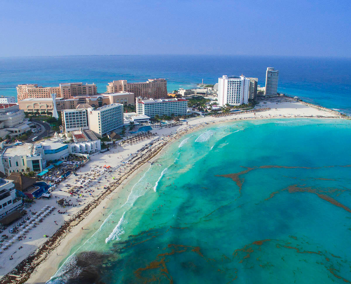 Vista aérea da Zona Hoteleira de Cancún, com areia branca, mar azul-turquesa e grandes hotéis alinhados à beira-mar.