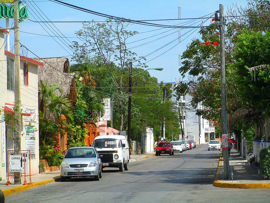 Rua residencial em Cancún, com árvores, carros estacionados e casas coloridas ao fundo.