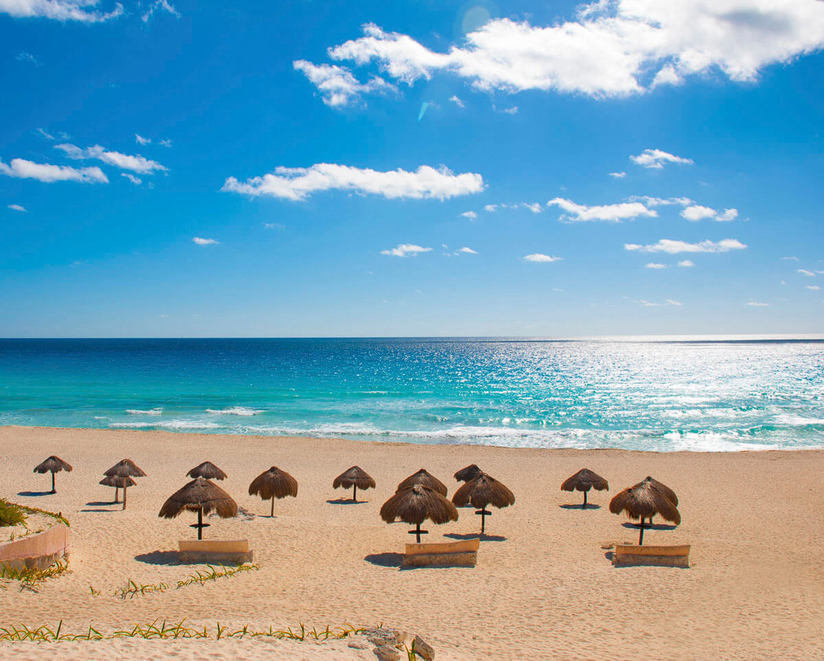 Praia de areia branca em Cancún com guarda-sóis de palha e mar azul cristalino ao fundo.