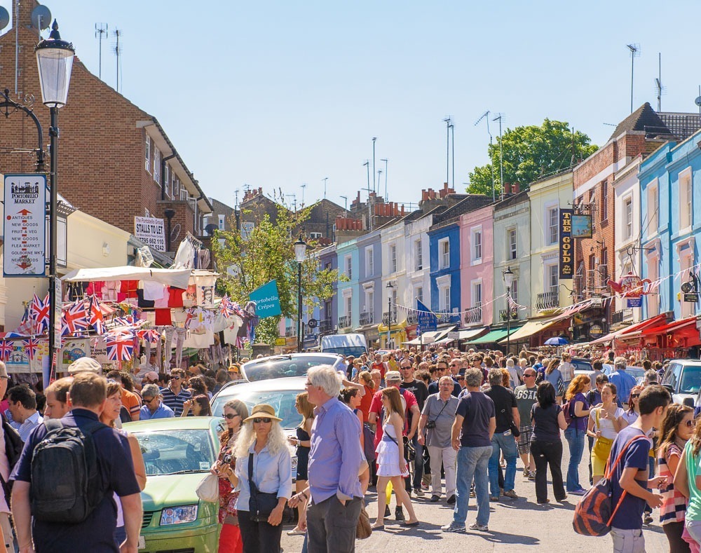 Mercado de Portobello Road em Notting Hill, com barracas coloridas, bandeiras do Reino Unido e uma grande multidão.