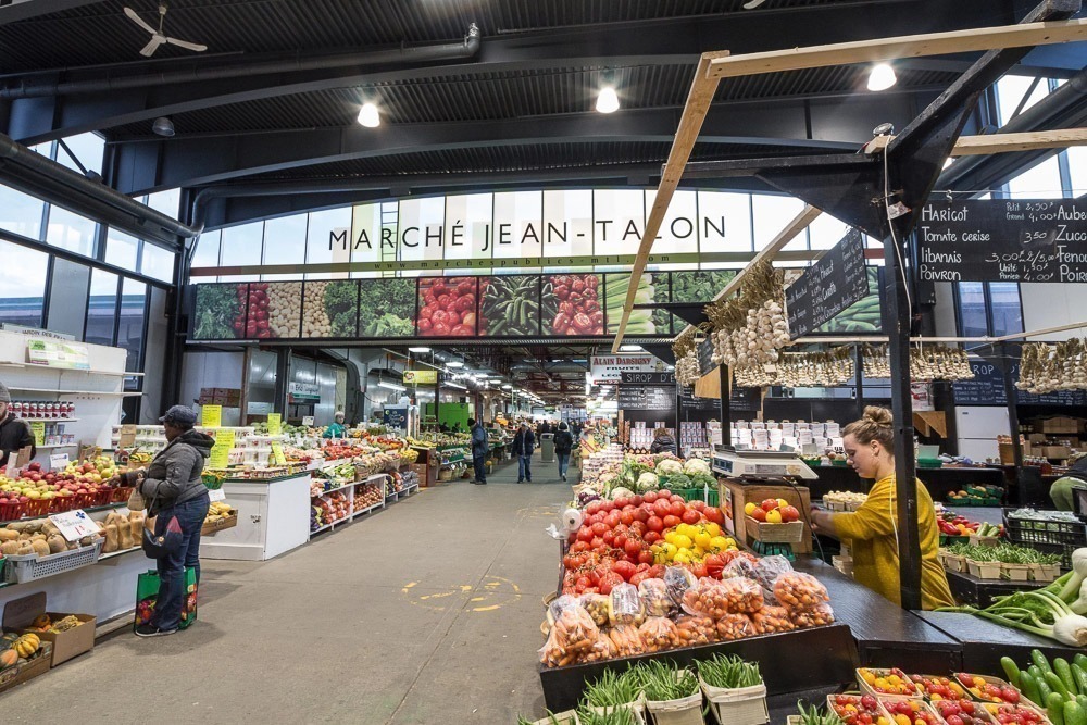 Mercado Jean-Talon em Montreal, com bancas de frutas, legumes e hortaliças frescas, além de clientes fazendo compras.