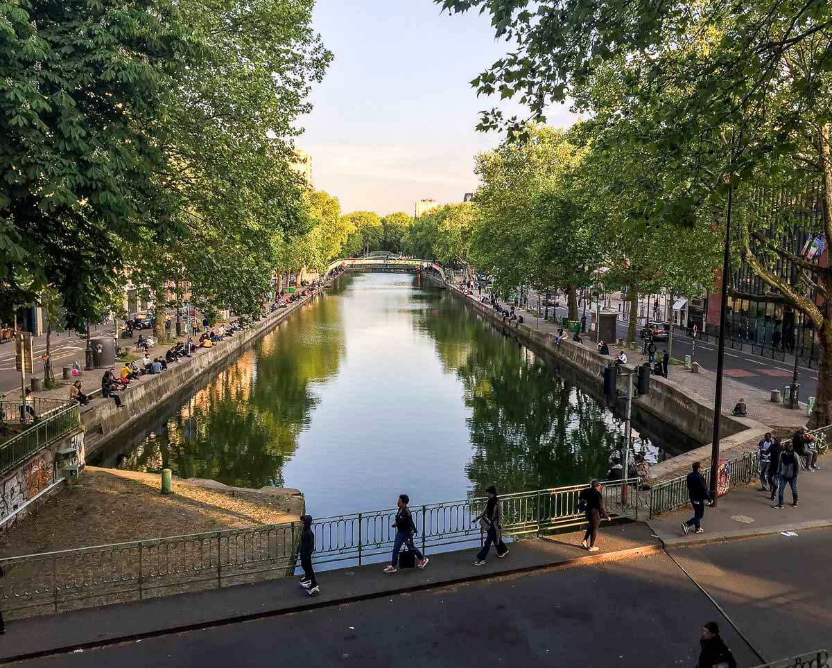 O Canal Saint-Martin, em Paris, cercado por árvores e calçadas movimentadas, com pessoas sentadas à beira da água e atravessando uma ponte.