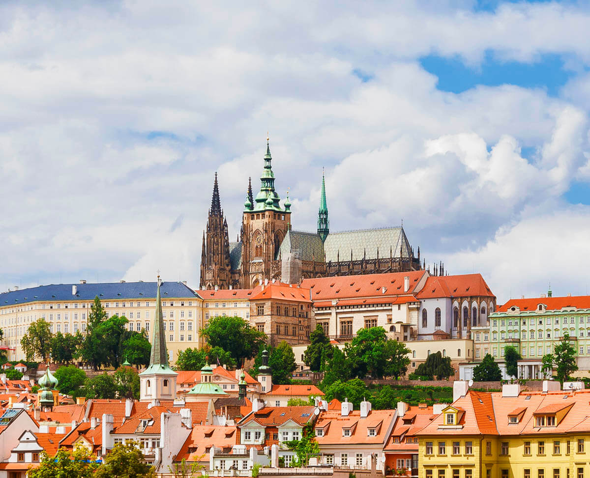 Vista ampla do Castelo de Praga com telhados alaranjados, torres góticas e arquitetura histórica cercada por vegetação.