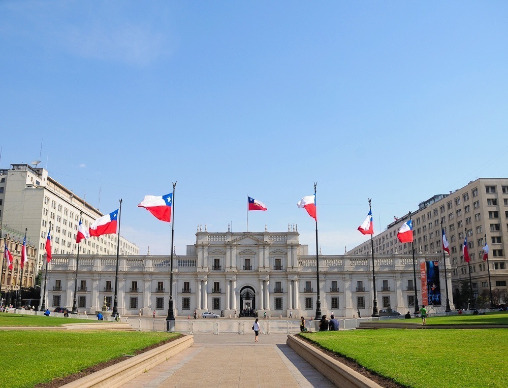 Fachada imponente do Palácio La Moneda, cercada por bandeiras chilenas e uma praça verde.