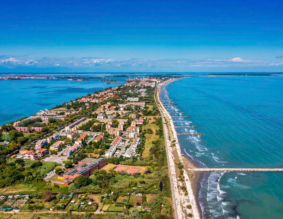 Vista aérea do Lido di Venezia, uma longa ilha entre a lagoa de Veneza e o mar Adriático, com praias, casas e vegetação.
