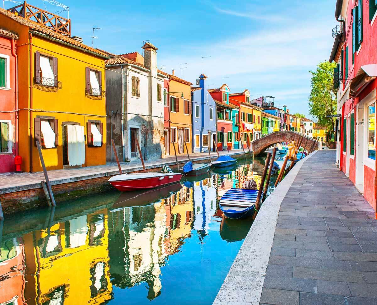 Canal de Burano, em Veneza, com casas coloridas refletindo na água e barcos atracados ao longo das margens.