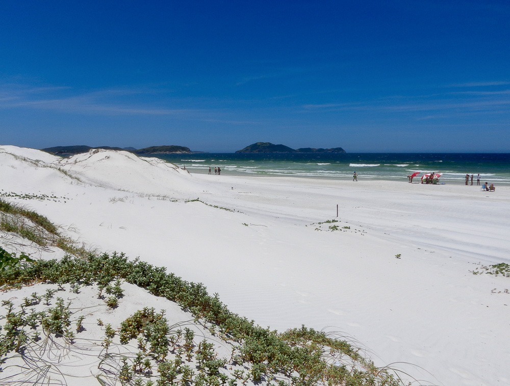 Areias brancas e vegetação rasteira na Praia das Dunas, com o oceano azul ao fundo.