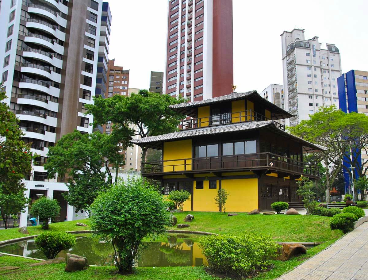 Memorial da Imigração Japonesa em Curitiba, um pagode amarelo e preto cercado por um jardim com lago e árvores, contrastando com os prédios modernos ao fundo.