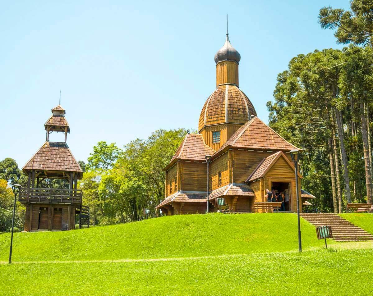 Memorial Ucraniano no Parque Tingui, com igreja de madeira e campanário cercados por um gramado verde e árvores.