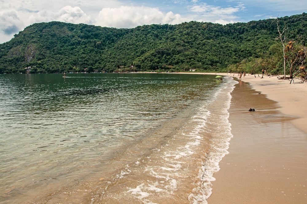 Praia tranquila em Paraty-Mirim, com mar calmo, areia dourada e montanhas cobertas por mata atlântica ao fundo.