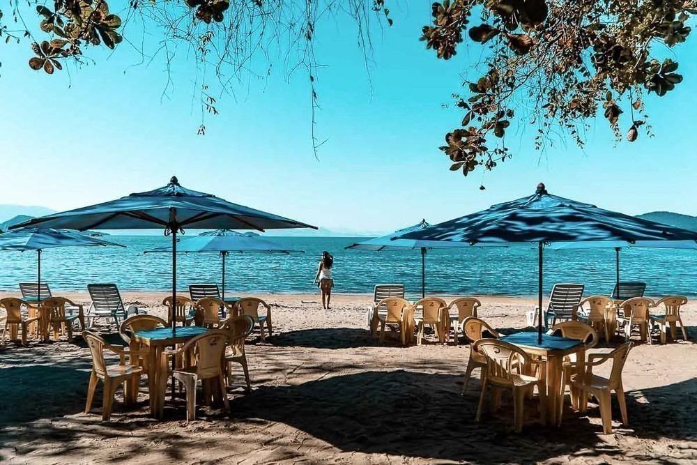 Praia do Pontal com mesas e cadeiras sob guarda-sóis azuis, à beira do mar calmo.