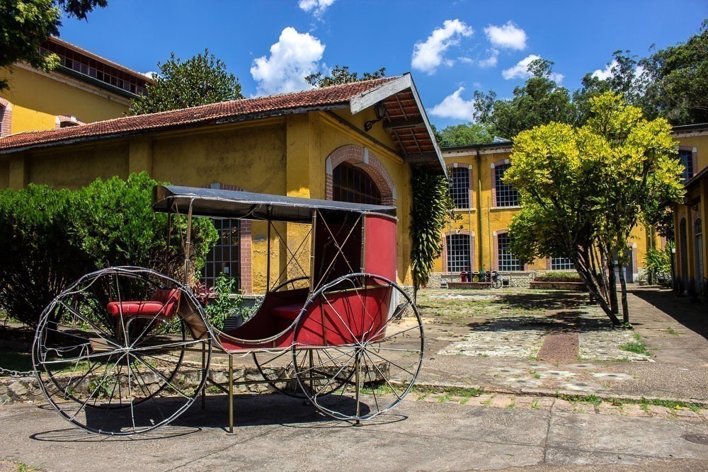 Carroça antiga vermelha com rodas de metal em frente a um edifício amarelo do Centro Cultural Brasital, com arquitetura histórica, rodeado por árvores e plantas.