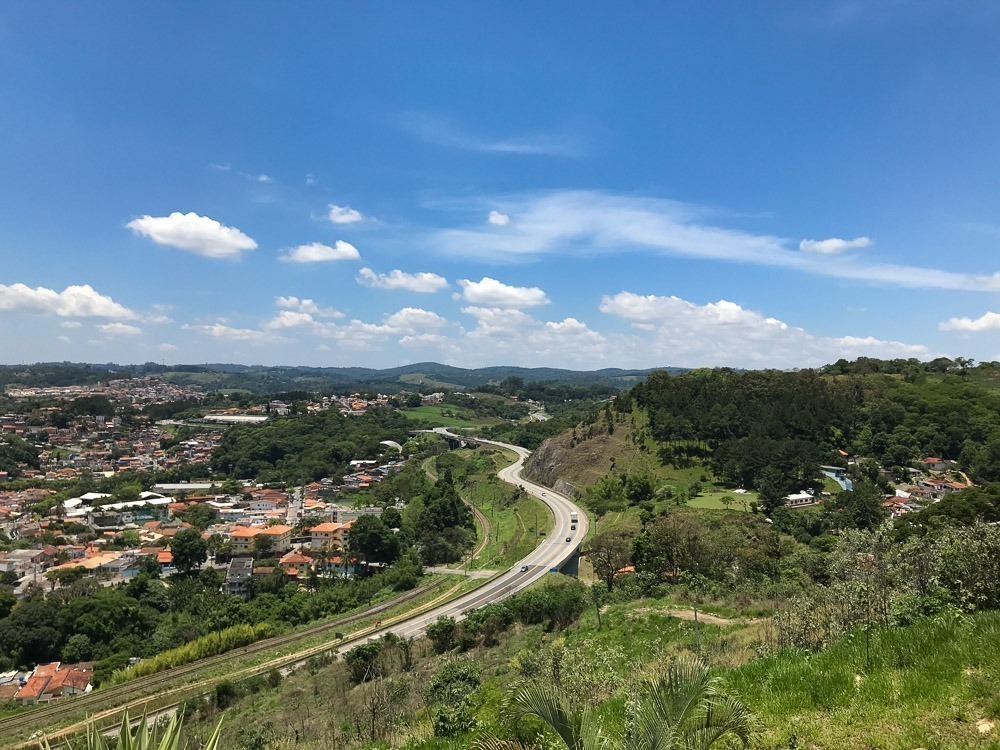 Estrada sinuosa cercada por vegetação e morros, ligando a cidade às montanhas, com céu azul e algumas nuvens.