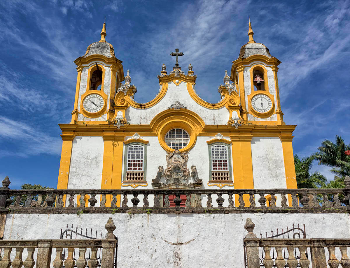 Fachada da Igreja Matriz de Santo Antônio, em Tiradentes, MG, com sua estrutura barroca em tons de branco e amarelo, com dois campanários e um céu azul ao fundo.