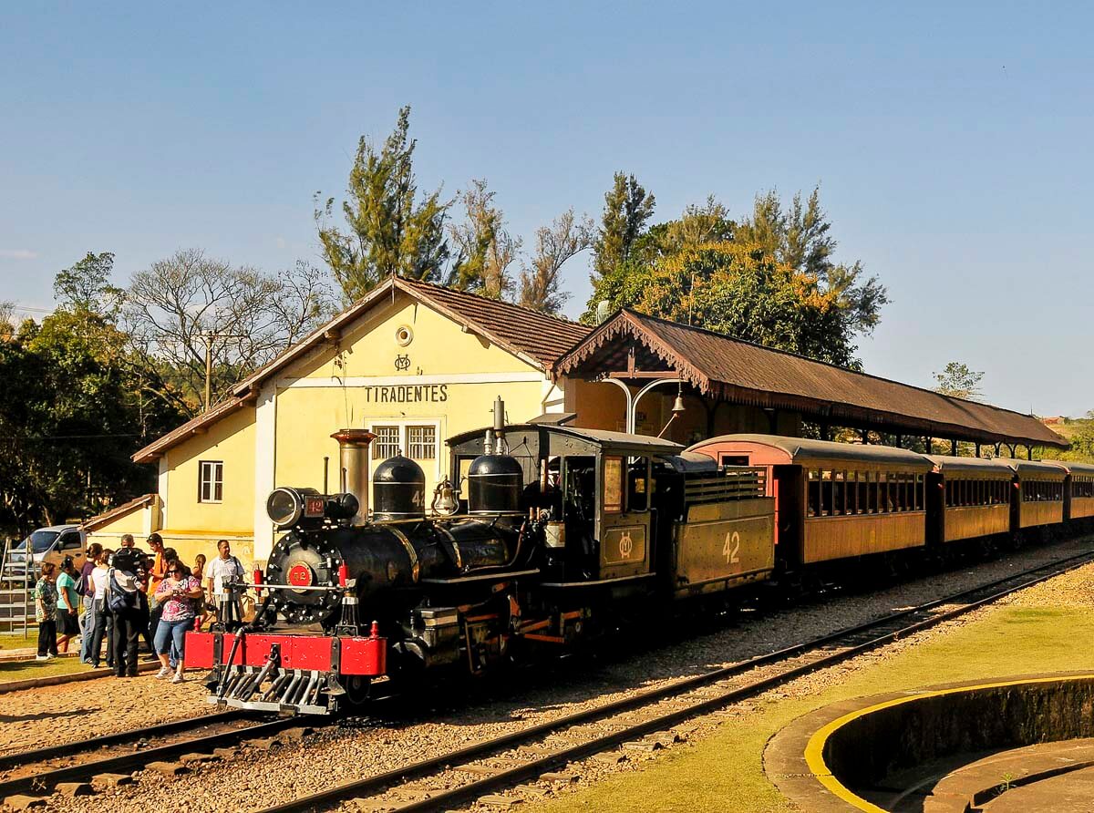 Trem Maria Fumaça estacionado na plataforma da estação ferroviária de Tiradentes, MG, com passageiros ao redor e um céu azul ao fundo.