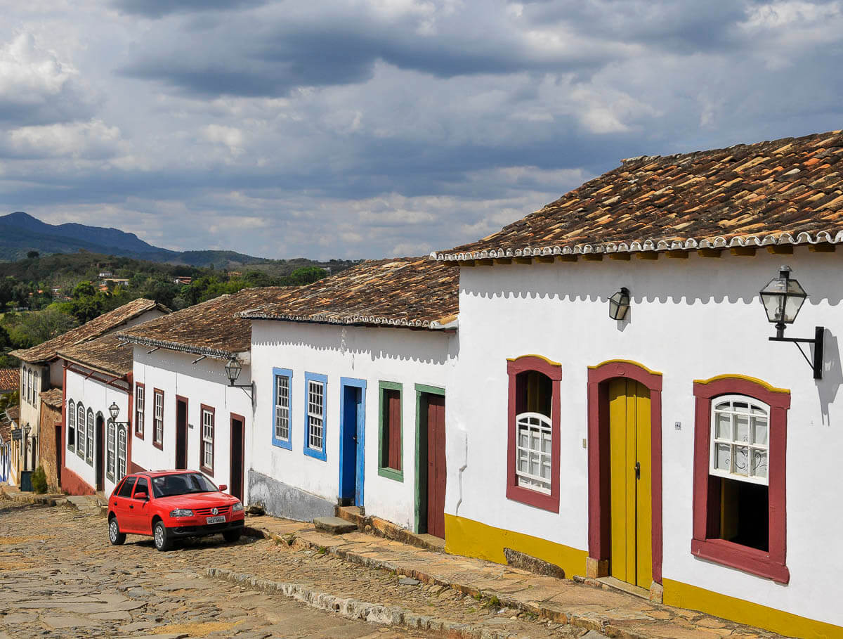 Rua de paralelepípedos no centro histórico de Tiradentes, MG, com casas coloniais brancas de portas e janelas coloridas e um carro vermelho estacionado.