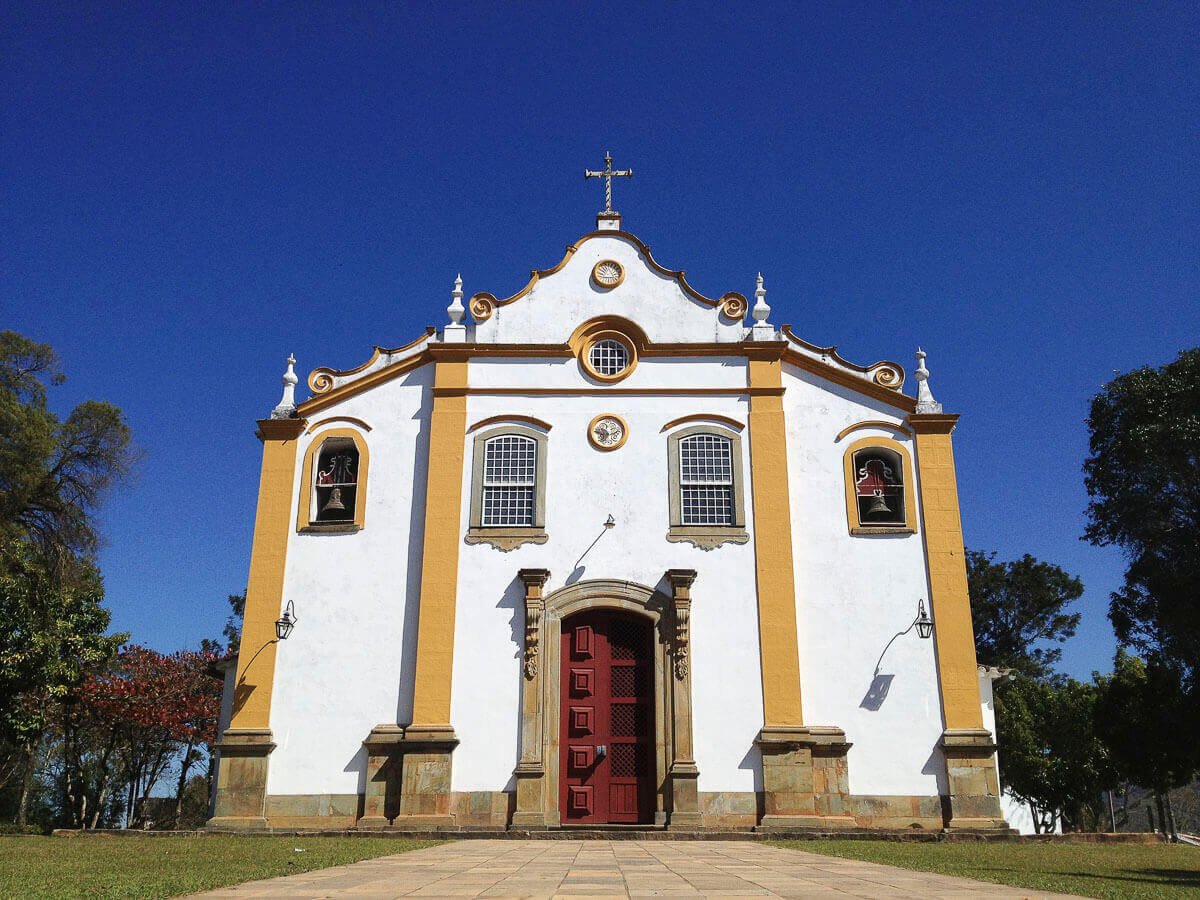 Fachada branca e amarela do Santuário da Santíssima Trindade, em Tiradentes, MG, com portas vermelhas e céu azul intenso ao fundo.